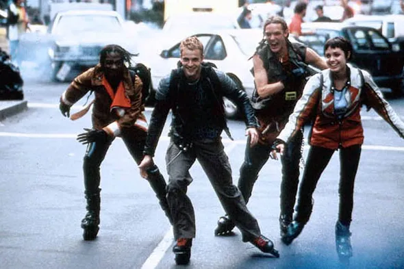 Still from Hackers (1995): four characters rollerblade toward the camera down a city street in 1990s club clothes.